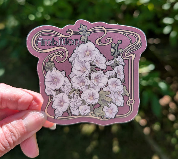 A hand holds a die-cut sticker featuring a  mauve background and yellow Art Nouveau design with the word 'Ambition' and a cluster of white hollyhock flowers, set against a blurred green outdoor background.