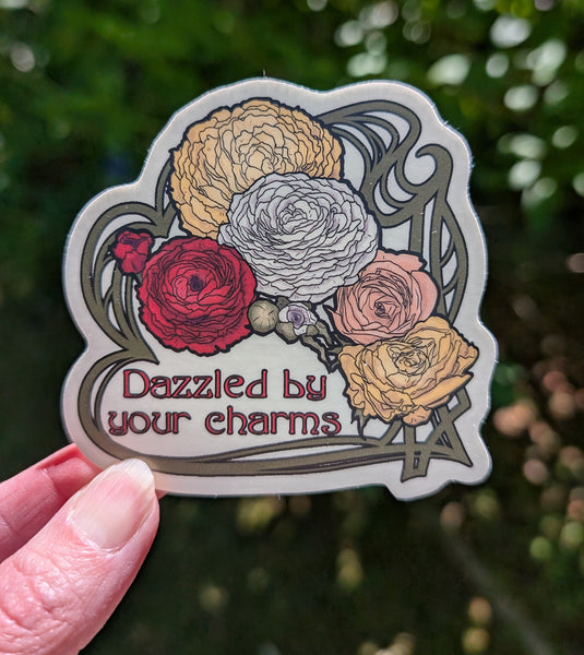A hand holds a die-cut sticker featuring an illustration of five ranunculus flowers in red, gold, peach, and white, with the text 'Dazzled by your charms' in dark red lettering. The background is a blurred green outdoor setting.
