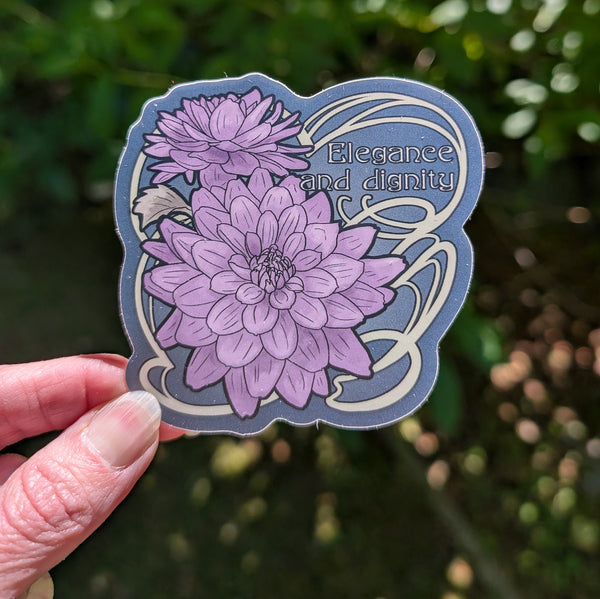 A hand holds a die-cut sticker featuring two purple dahlias on a slate blue background with white Art Nouveau-style swirls and the words 'Elegance and dignity.' The background is a blurred outdoor green garden.
