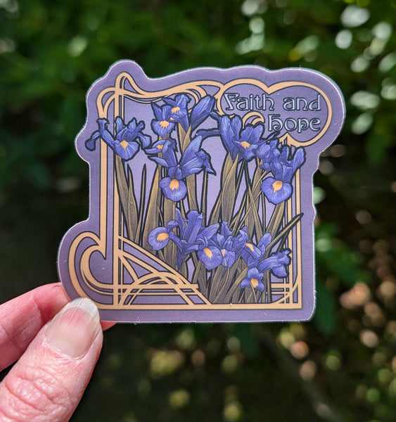 A hand holds a die-cut sticker featuring blue irises on a dark lavender background and the words 'Faith and hope' in a gold Art Nouveau frame, with a blurred green outdoor background.