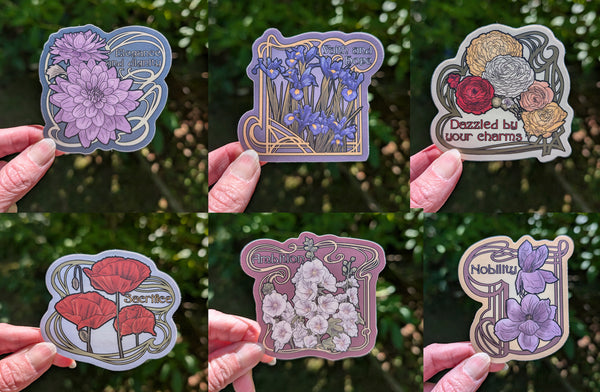 Six photos, each a hand holding a sticker with a flower, an art nouveau-styled border, and meaning of the flower with blurred green outdoor background. Please see individual alt text for each flower and meaning.