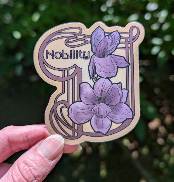 A hand holds a die-cut sticker featuring pale purple magnolias on a gold background and the word 'Nobility' in an Art Nouveau style, with a blurred green outdoor background.