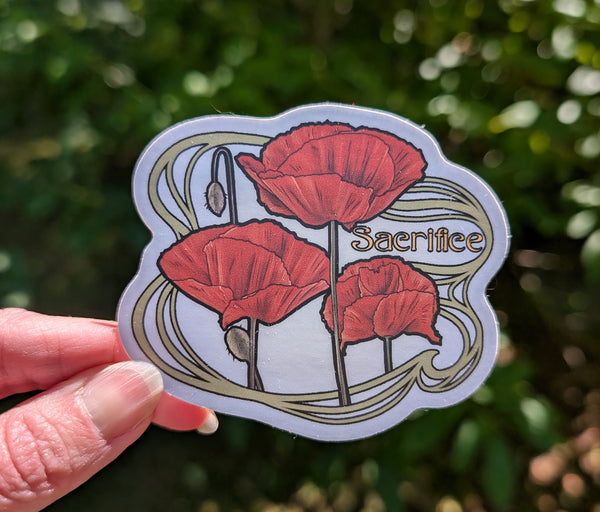 A hand holds a die-cut sticker featuring three red poppies, a seed pod, and the word 'Sacrifice' in gold lettering, set against a blurred green outdoor background.