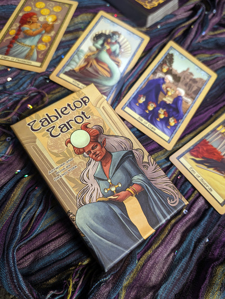 The Tabletop Tarot deck box, with four cards above, laying on a striped scarf.